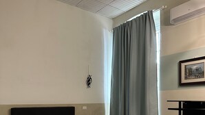Habitación de hotel con cama, escritorio, silla, ventana con cortinas y unidad de aire acondicionado.