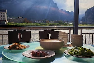 A table with a bowl of soup, a plate of vegetables, and a plate of meat, set against a scenic river view.