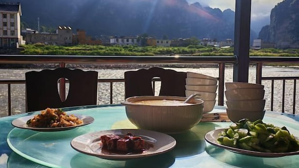 Een tafel met een kom soep, een bord vlees en een bord groenten, met op de achtergrond een mooi rivierzicht en bergen.