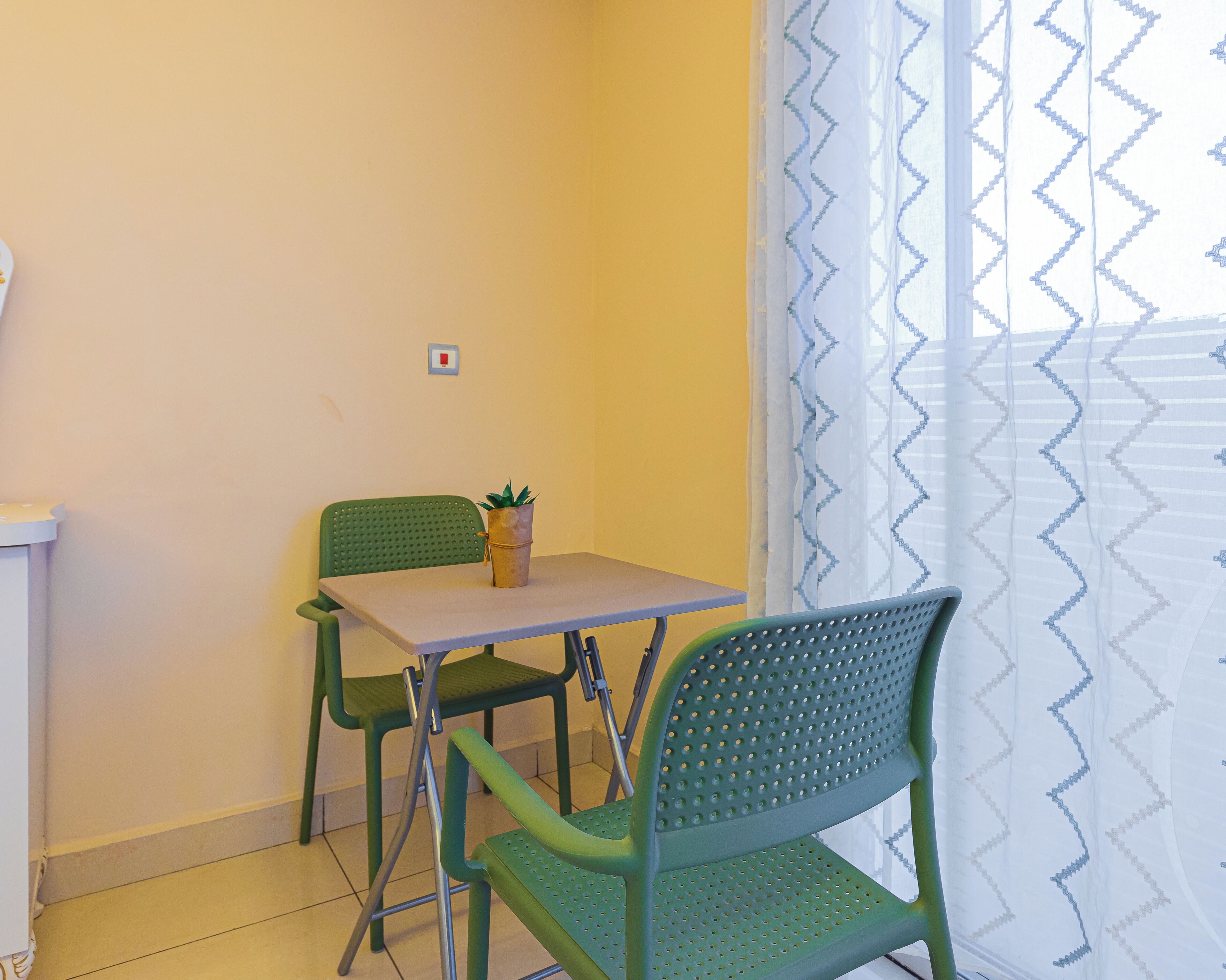 Une petite pièce avec une table et deux chaises, une plante en pot sur la table et une fenêtre ornée d’un rideau à motifs.