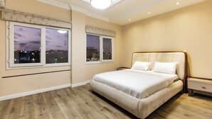 Un dormitorio con una cama grande, dos mesitas de noche y una ventana con vistas a la ciudad al anochecer.