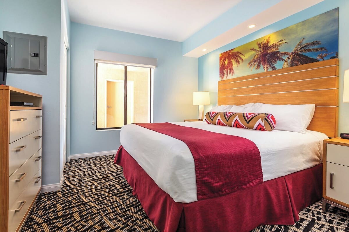 Quarto de hotel com cama, mesa de cabeceira, luminária, quadro de palmeiras, janela e televisão fixada na parede.