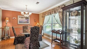 Un coin repas comprenant une table et des chaises, un lustre, une vitrine contenant des objets décoratifs et une fenêtre avec des rideaux.