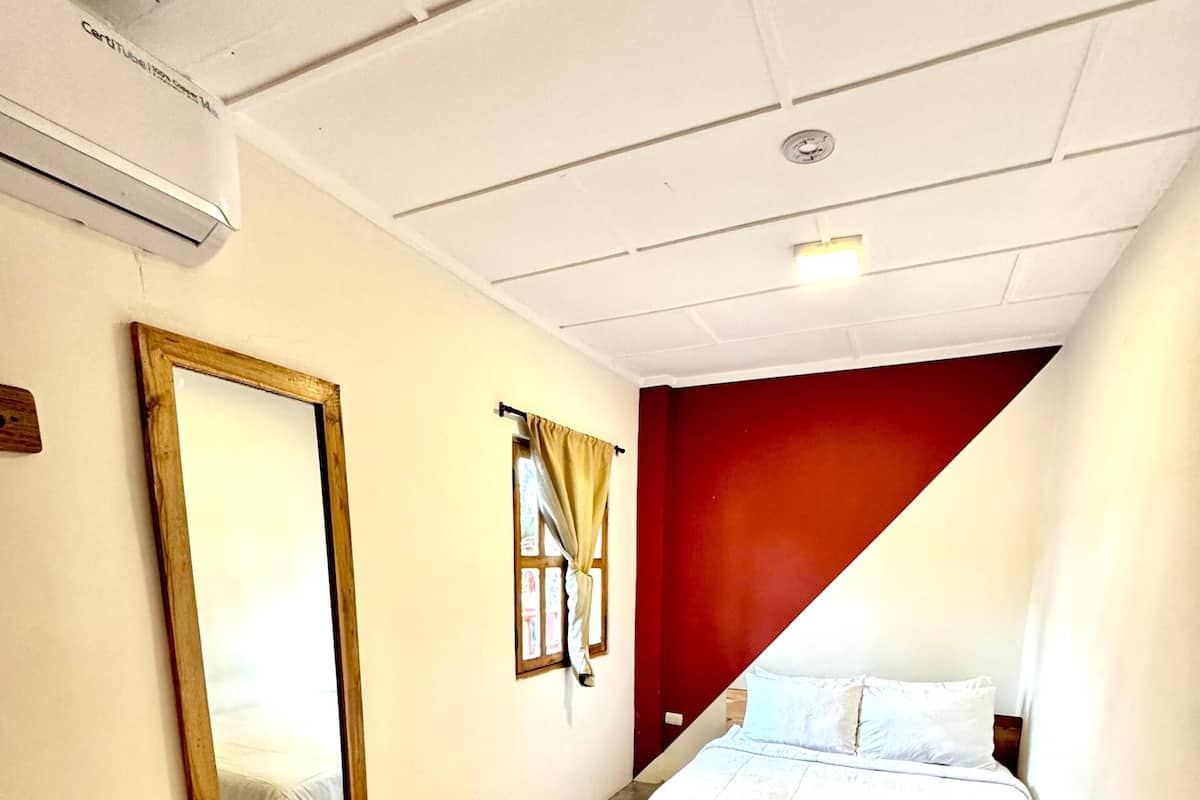 A hotel room with a bed, a small table with a towel, a mirror, and a window with curtains.