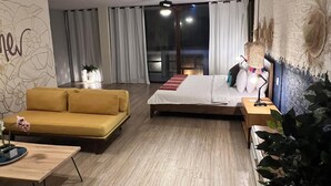 A modern hotel room with a wooden floor, a bed with a red and white striped bedspread, a yellow sofa, a small table with a lamp, and a large window with curtains. - Hotel Montañita (Montañita)