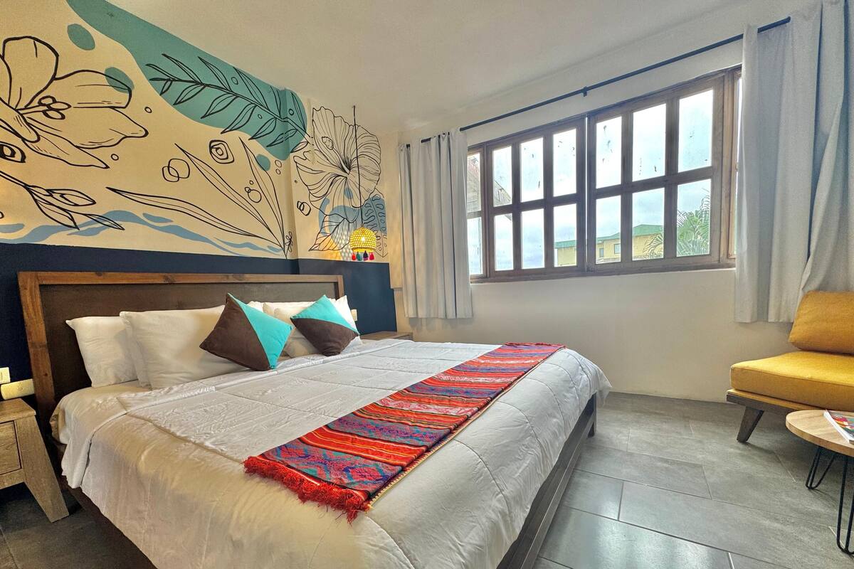 A neatly made bed with a white comforter and a colorful throw blanket, a wooden headboard, a yellow armchair, and a large window with curtains.