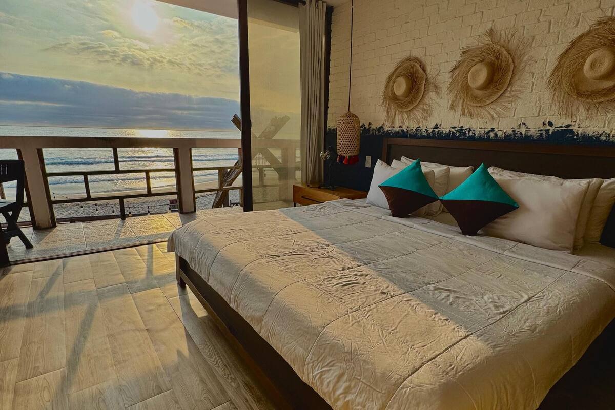 A bedroom with a large bed, a ceiling fan, a view of the ocean, and a brick wall with decorative hats.