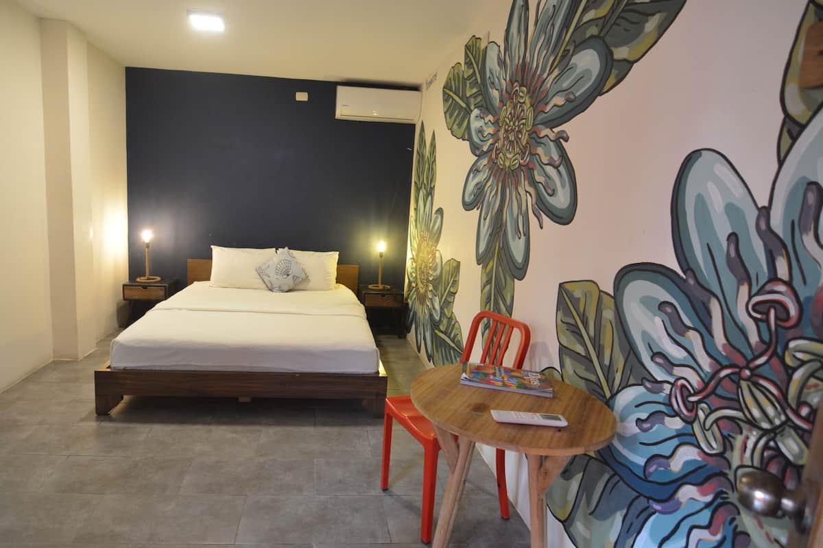 A bedroom with a bed, a nightstand, a table with a chair, and a wall with a floral mural.