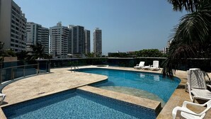 Une piscine sur le toit, avec une eau d’un bleu cristallin, entourée de chaises longues blanches et d’un palmier.