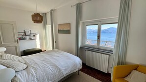 Un dormitorio con cama, un sillón amarillo, un ventanal con vista al mar, un radiador y una lámpara colgante.
