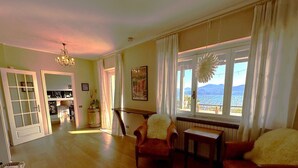 Un salón espacioso con suelo de madera, un candelabro, una zona de comedor con mesa y sillas, y un ventanal con vistas al exterior.