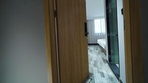 Un couloir d’hôtel avec une porte en bois portant le numéro « 8121 », qui donne accès à une chambre équipée d’un lit et séparée par une cloison en verre.