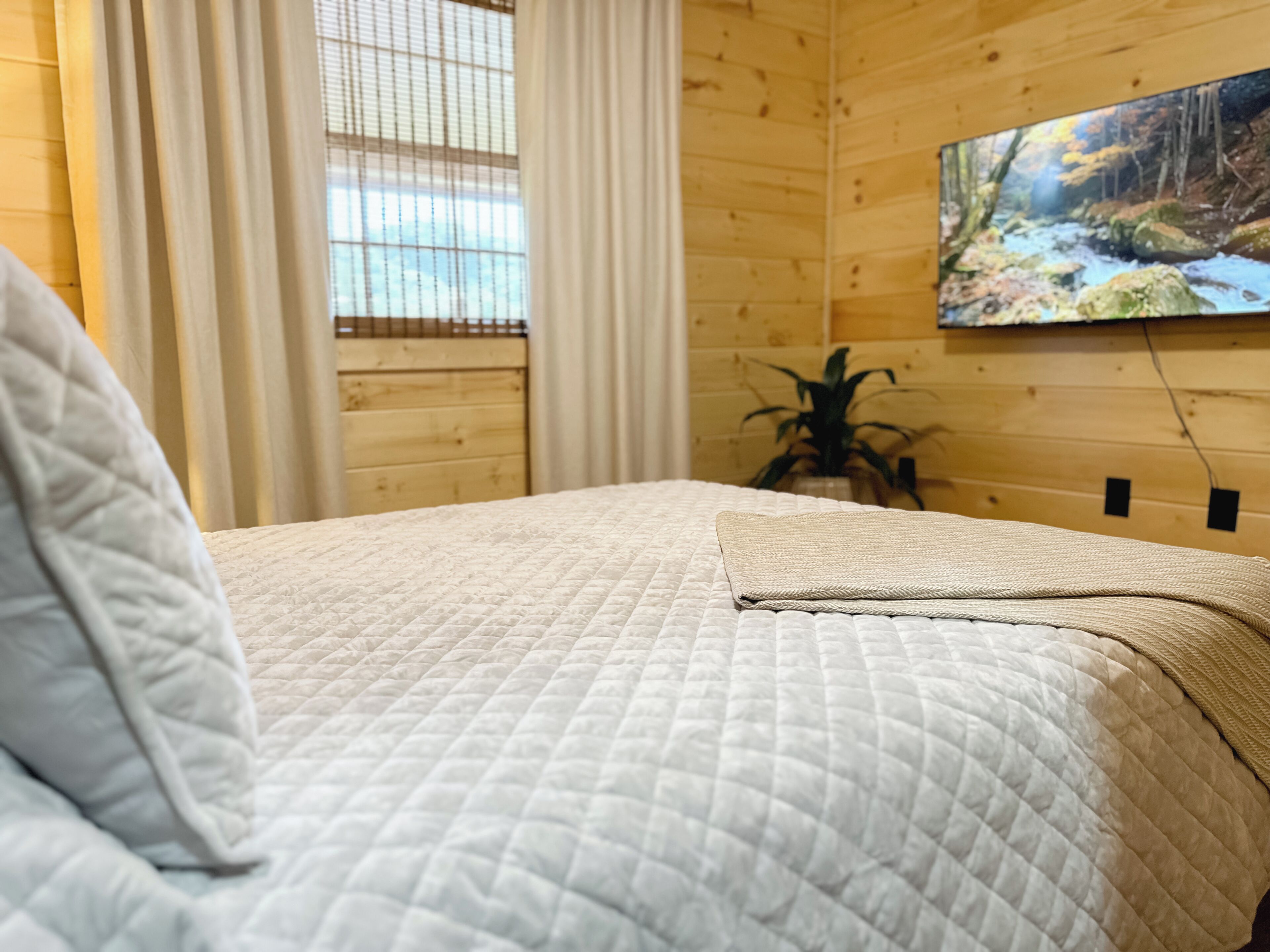 Una habitación de cabaña de madera con una cama, una ventana con cortinas y un cuadro de un arroyo en el bosque.
