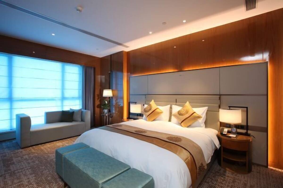 Une chambre d’hôtel moderne avec un grand lit, un canapé, une petite table et une fenêtre avec des stores.