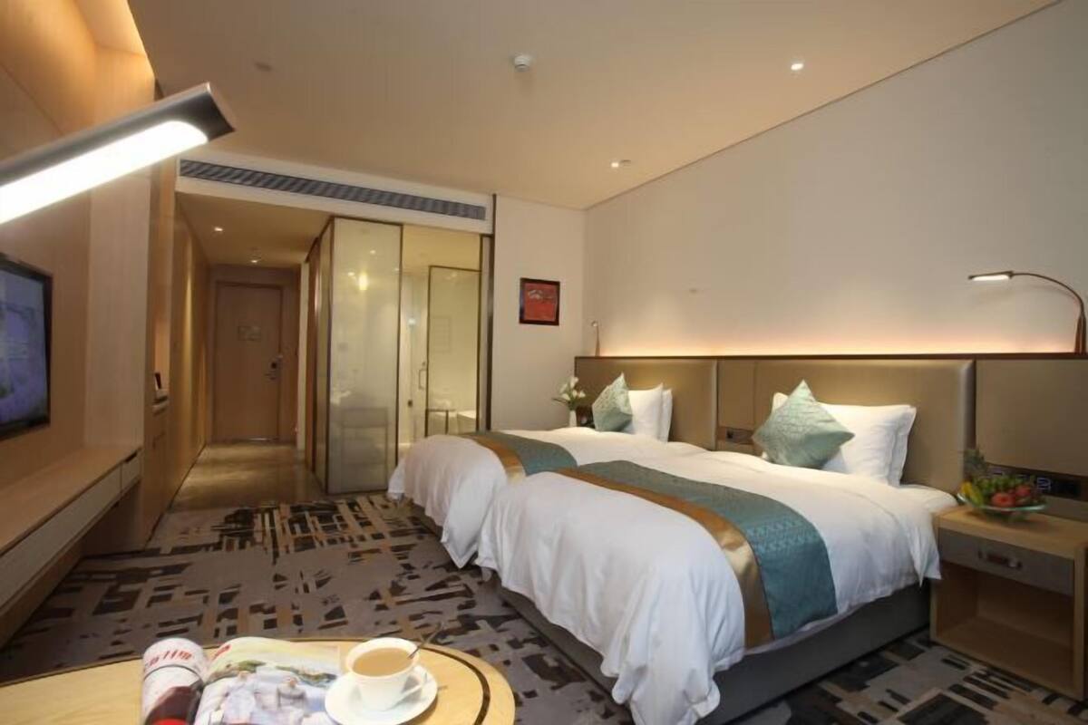 Une chambre d’hôtel avec deux lits, une télévision, une table basse avec une tasse et une table de chevet avec un panier de fruits.