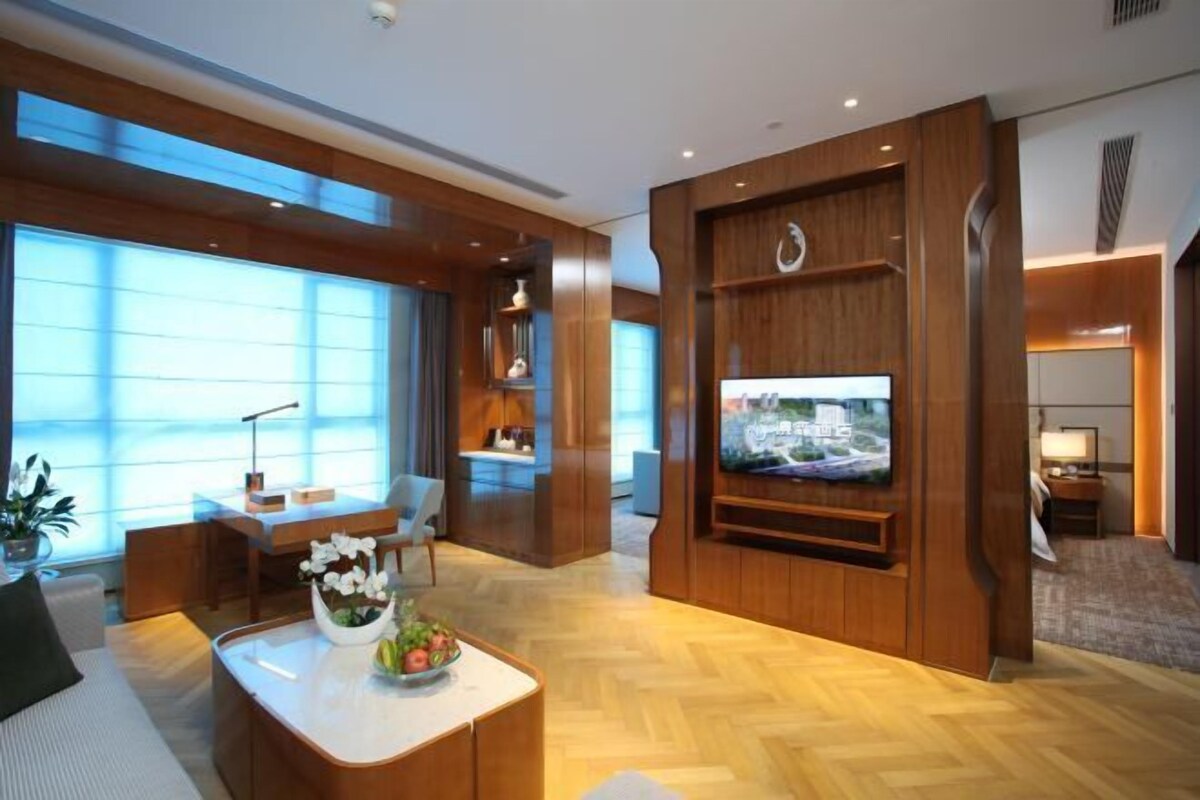 A modern living room with a flat-screen TV, wooden furniture, and large windows.