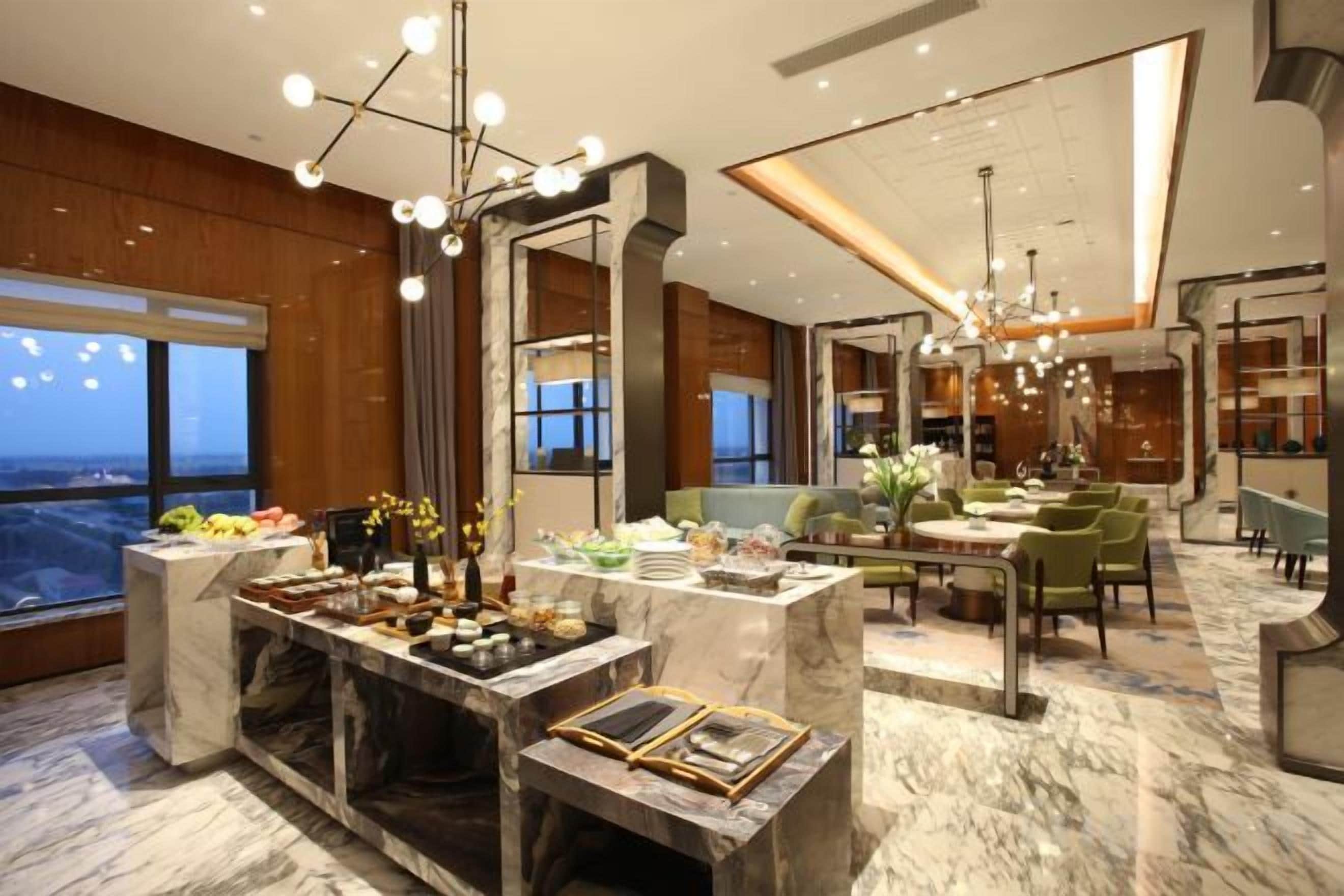 A modern hotel lobby with a marble reception desk, a dining area with green chairs, and a large window offering a city view.