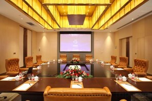 Ein Konferenzraum mit langem Tisch, mehreren Stühlen, einer Projektionsleinwand und einer Blumenarragem.