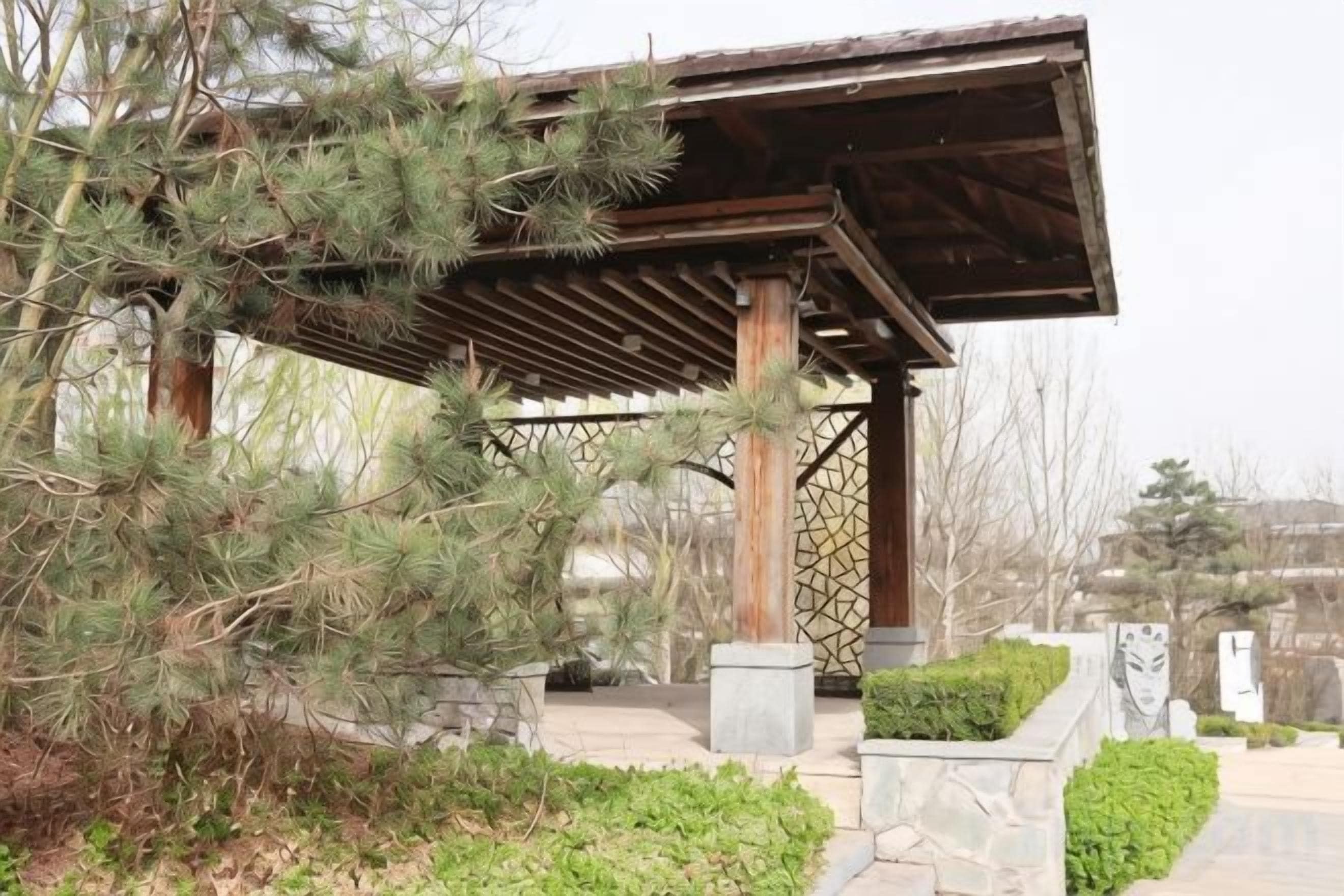 A wooden pavilion with a roof, surrounded by greenery and stone elements.