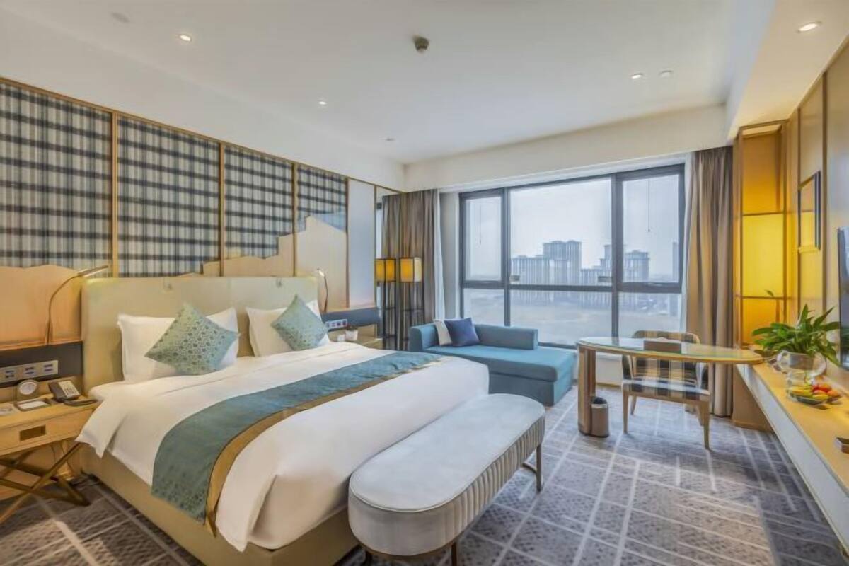 Une chambre d’hôtel avec un grand lit, un canapé, une petite table et une vue sur la ville grâce à de grandes fenêtres.