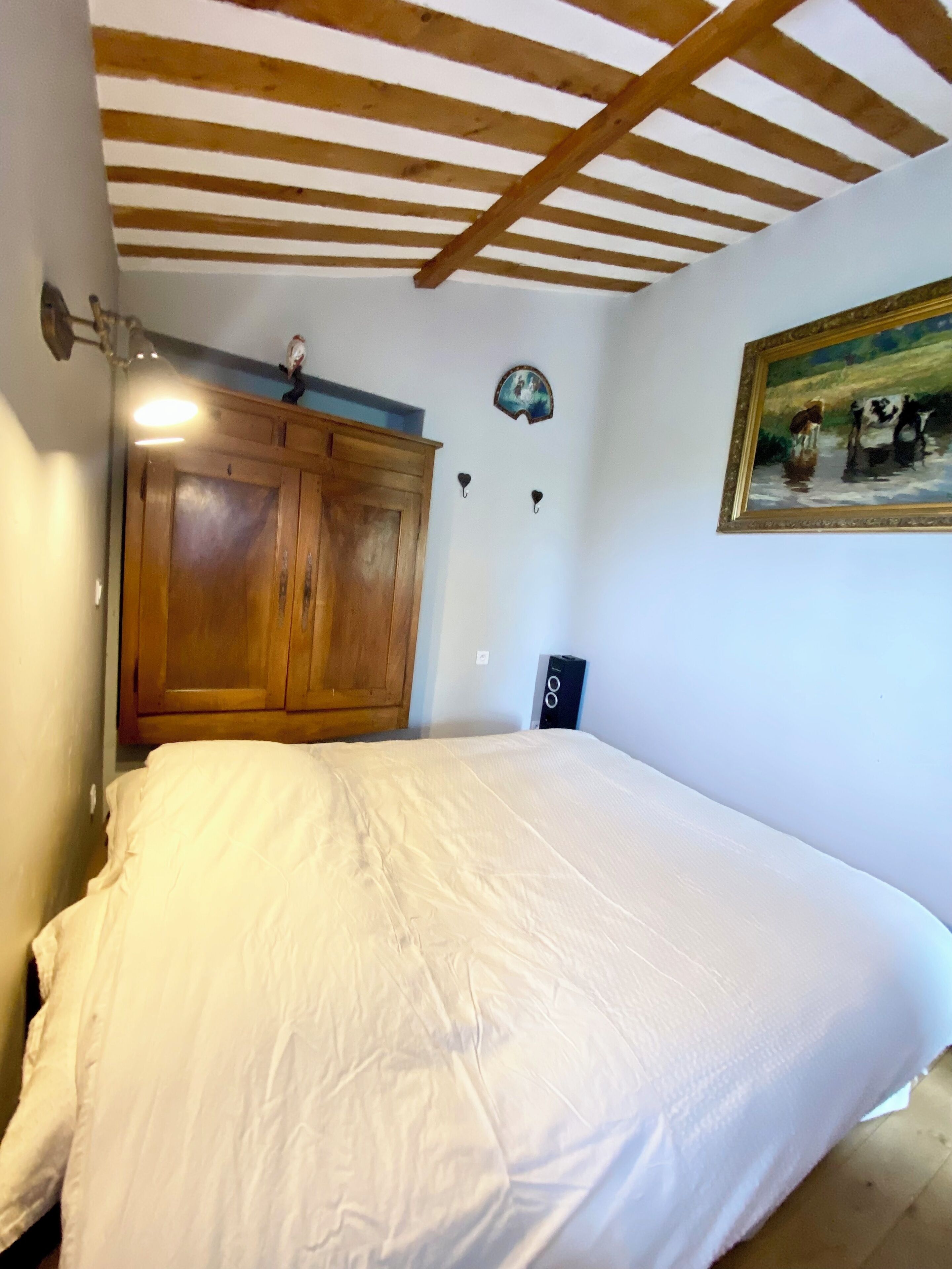 A bedroom with a wooden ceiling, a large bed, a wooden wardrobe, a painting on the wall, and a small table with a lamp.