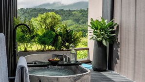 Une salle de bain extérieure moderne, dotée d’une grande baignoire, d’un porte-serviettes et offrant une vue sur une végétation luxuriante et des montagnes.