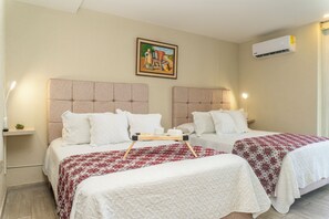 Ein Hotelzimmer mit zwei Betten, jeweils mit weißem Kopfteil und einer Bettdecke in Rot und Weiß. Darüber hängt ein Gemälde an der Wand, und eine Klimaanlage ist an der Wand montiert.