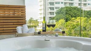 Een hottub met een houten bank en handdoeken, een fles wijn en glazen op een balkon met uitzicht op een stadsbeeld en groen.