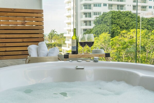 A hot tub with a wooden bench and towels, a bottle of wine, and glasses on a balcony overlooking a cityscape and greenery.