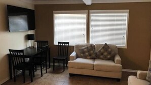 Ein Wohnzimmer mit einem beigefarbenen Sofa, einem Couchtisch und einem Essbereich mit Tisch und Stühlen. Zwei Fenster mit Jalousien sind vorhanden.