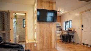 A modern hotel room with a wooden floor, a flat-screen TV mounted on a wall, a small kitchen area with a microwave and a dining table, and a wooden door with a vertical slat design.