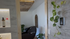 A modern room with a wooden ceiling, a massage chair, a small potted plant, and a wall-mounted shelf with bottles.