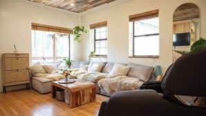A living room with a sofa, a coffee table, and a TV. There are windows and a ceiling with wooden beams.