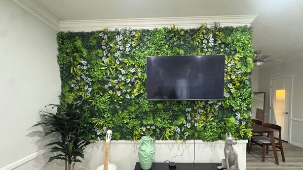 Ein Wohnzimmer mit an einer grünen, blätterreichen Wand montiertem Fernseher, einer Gitarre, einer Statue und einer kleinen Pflanze.