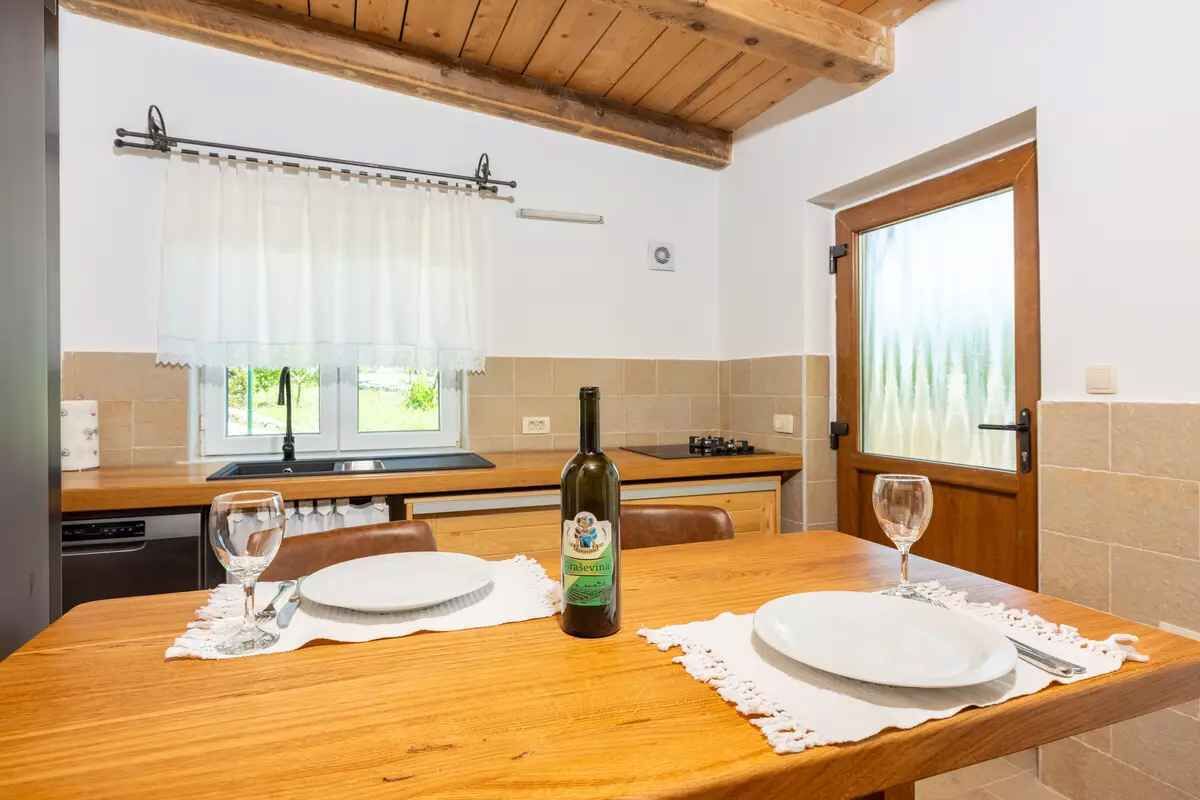 Une table à manger en bois, avec de l’assiette blanche et une bouteille de vin, une fenêtre avec un rideau et un coin cuisine avec un évier et une cuisinière.