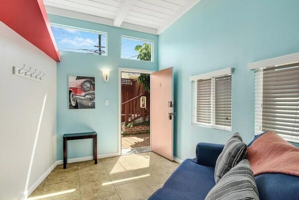 Ein Zimmer mit blauer Wand, einer Tür, die nach draußen führt, einem kleinen Tisch und einem blauen Sofa mit Kissen.