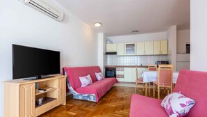 A compact living space with a TV on a wooden cabinet, a pink sofa, and a kitchen area with cabinets and a dining table.