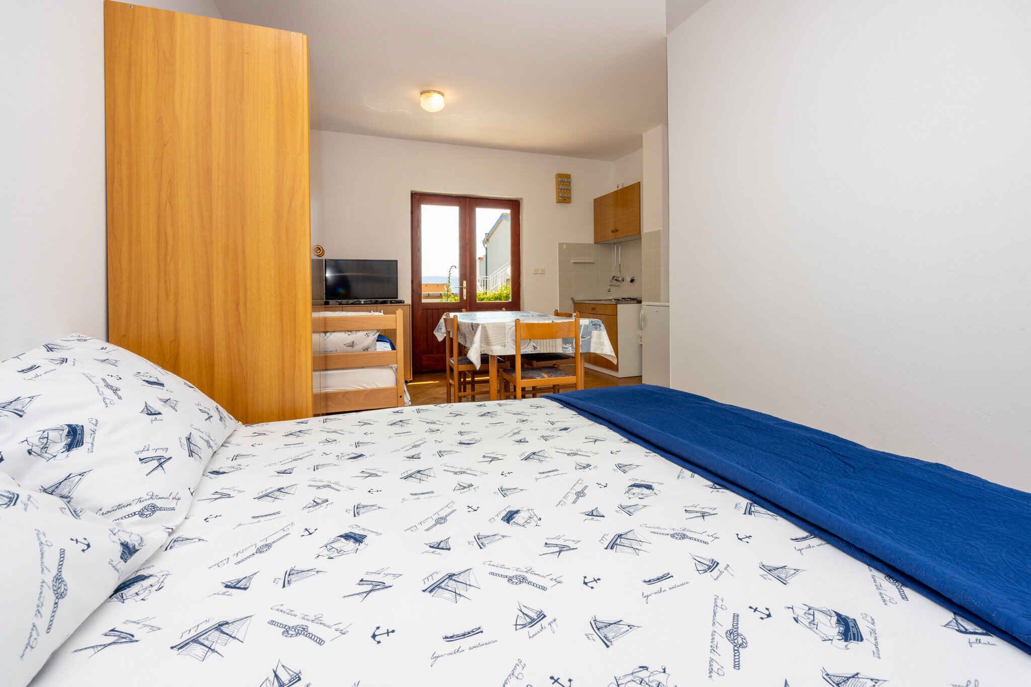 Un dormitorio con cama, un armario de madera, un escritorio con silla, televisor y una zona de cocina.