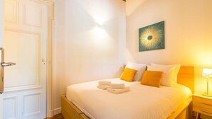 Una cama bien hecha con almohadas blancas y amarillas, una lámpara de noche y una obra de arte colgada en la pared.
