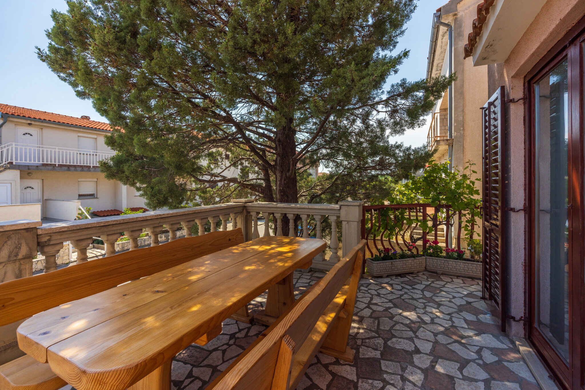 A wooden bench on a stone patio with a large tree and a building in the background.