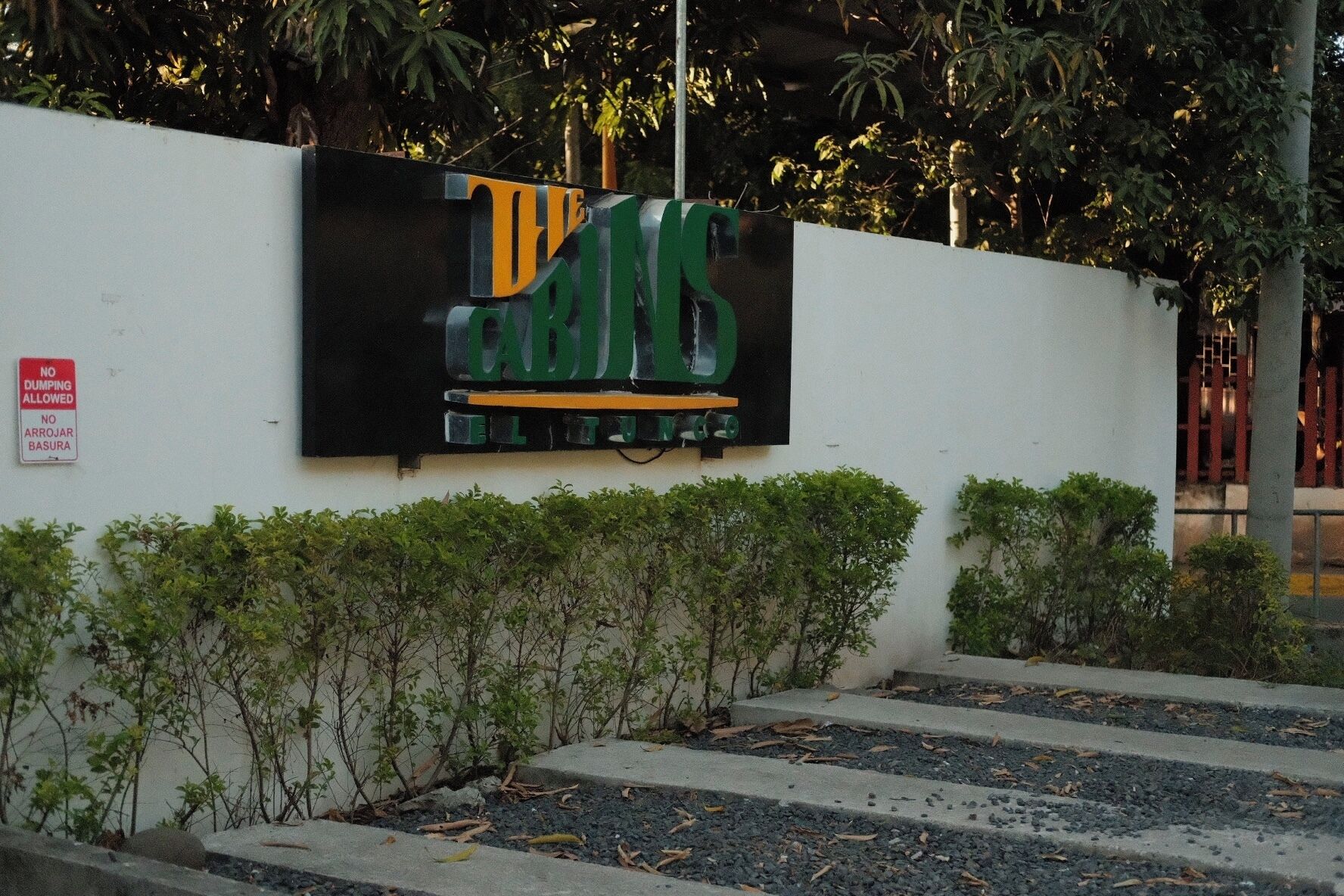 A sign with the text "THE GRINS" on a white wall, surrounded by greenery and a red sign prohibiting dumping.