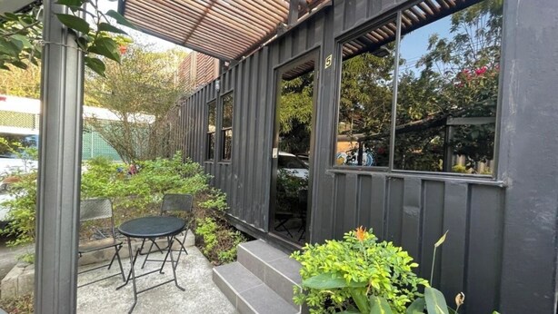 Uma área de estar coberta ao ar livre, com mesa e cadeiras, rodeada por plantas e uma pequena escadaria que leva a uma porta.