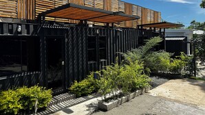 Um edifício moderno com fachada de ripas de madeira, área de estar coberta ao ar livre e um jardim paisagístico com diversos arbustos.