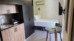 Quarto de hotel pequeno com cozinha compacta, uma cama e uma mesinha.