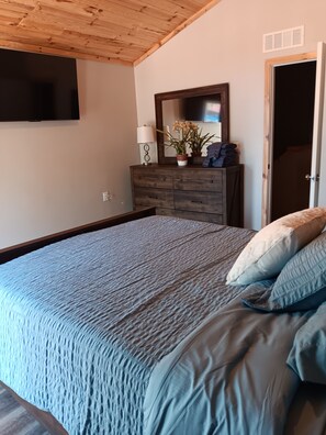 A bedroom with a wooden ceiling, a bed with a blue quilt, a dresser with a mirror, and a television mounted on the wall.