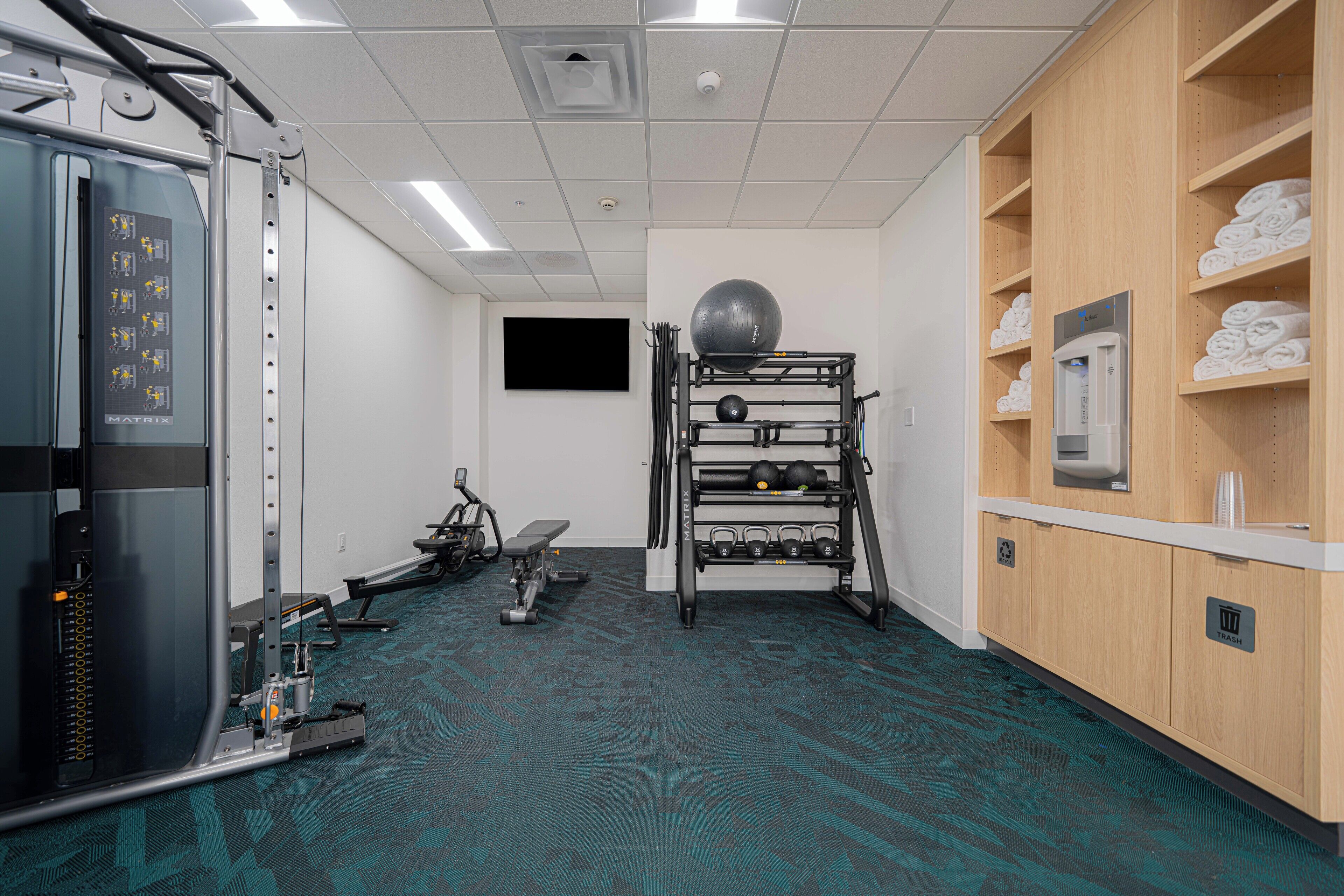 A gym with exercise equipment, a wall-mounted TV, and a rack with towels.