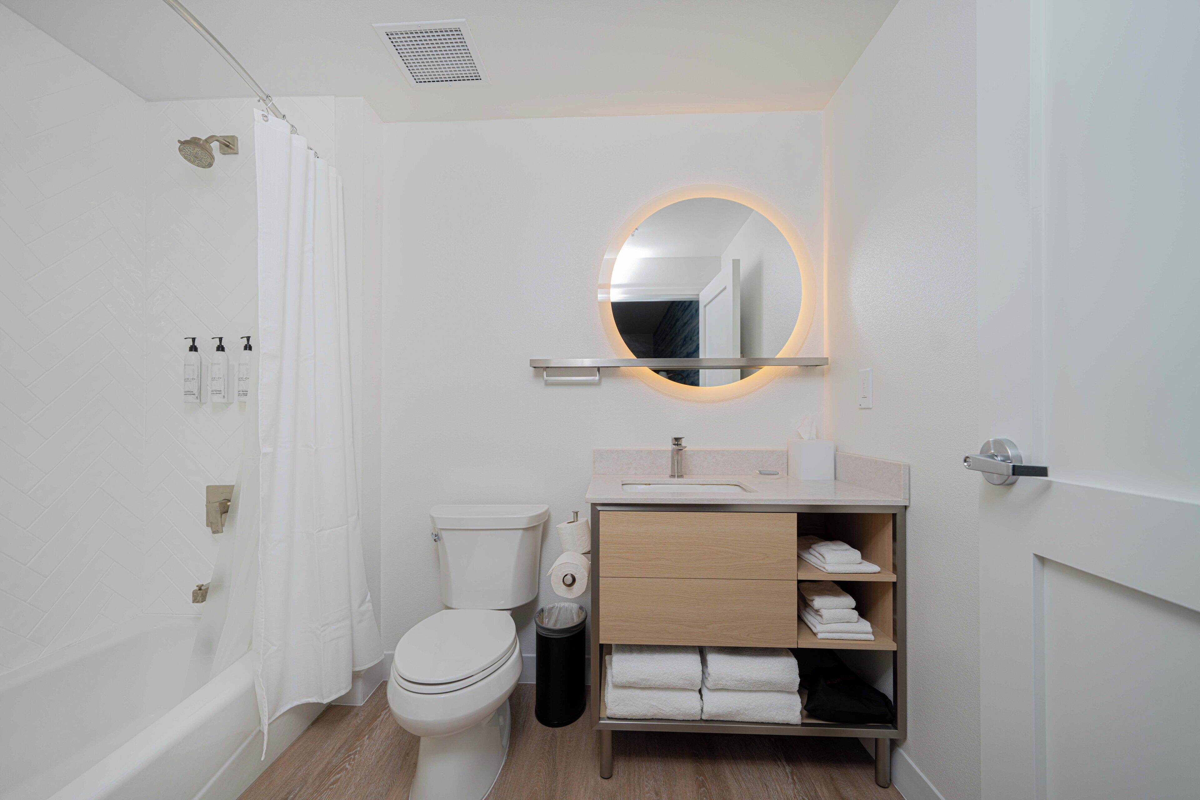 A modern bathroom with a toilet, shower, vanity, and a mirror with a circular light.