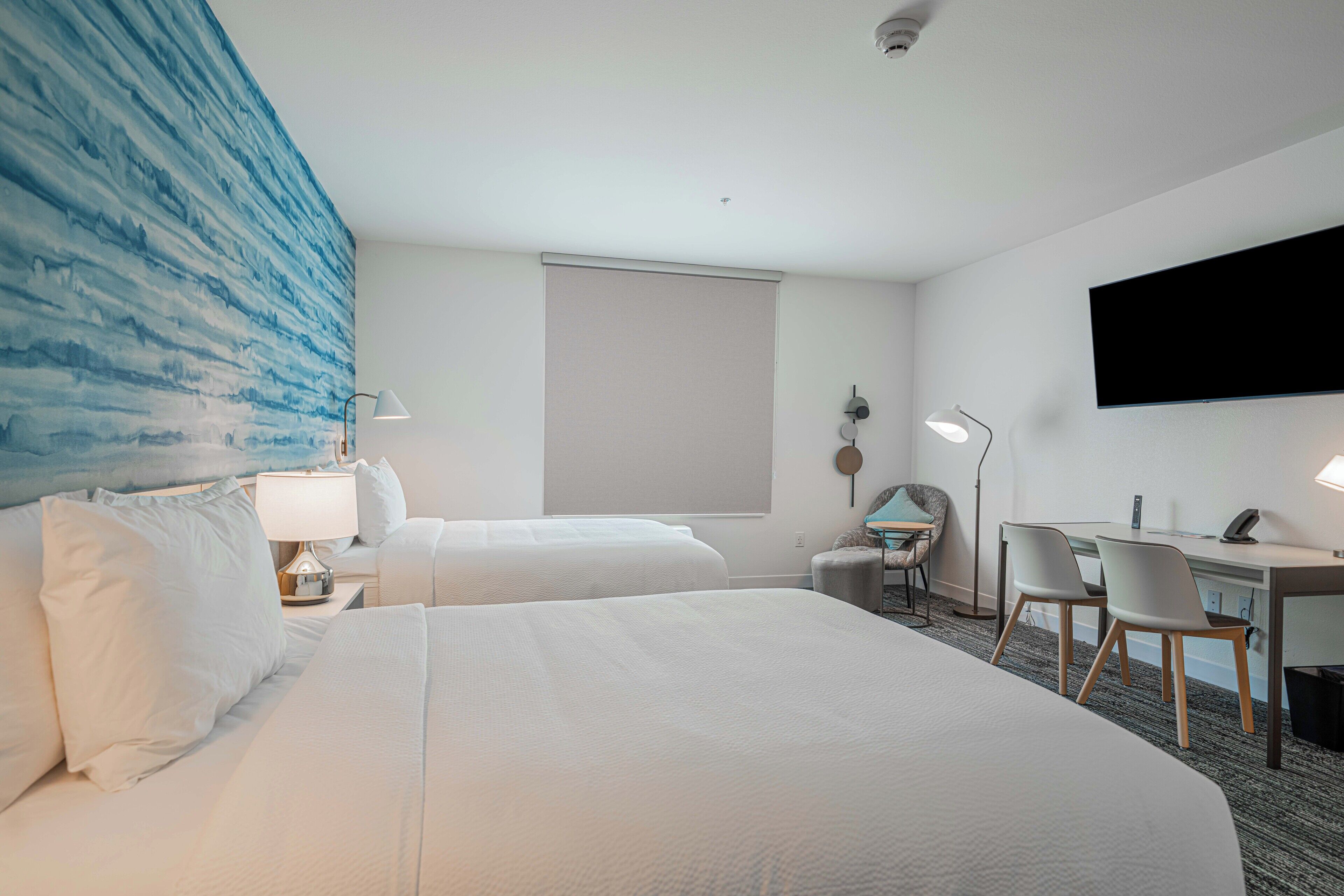 Habitación de hotel con dos camas, un escritorio, sillas, un televisor y una obra de arte azul grande en la pared.