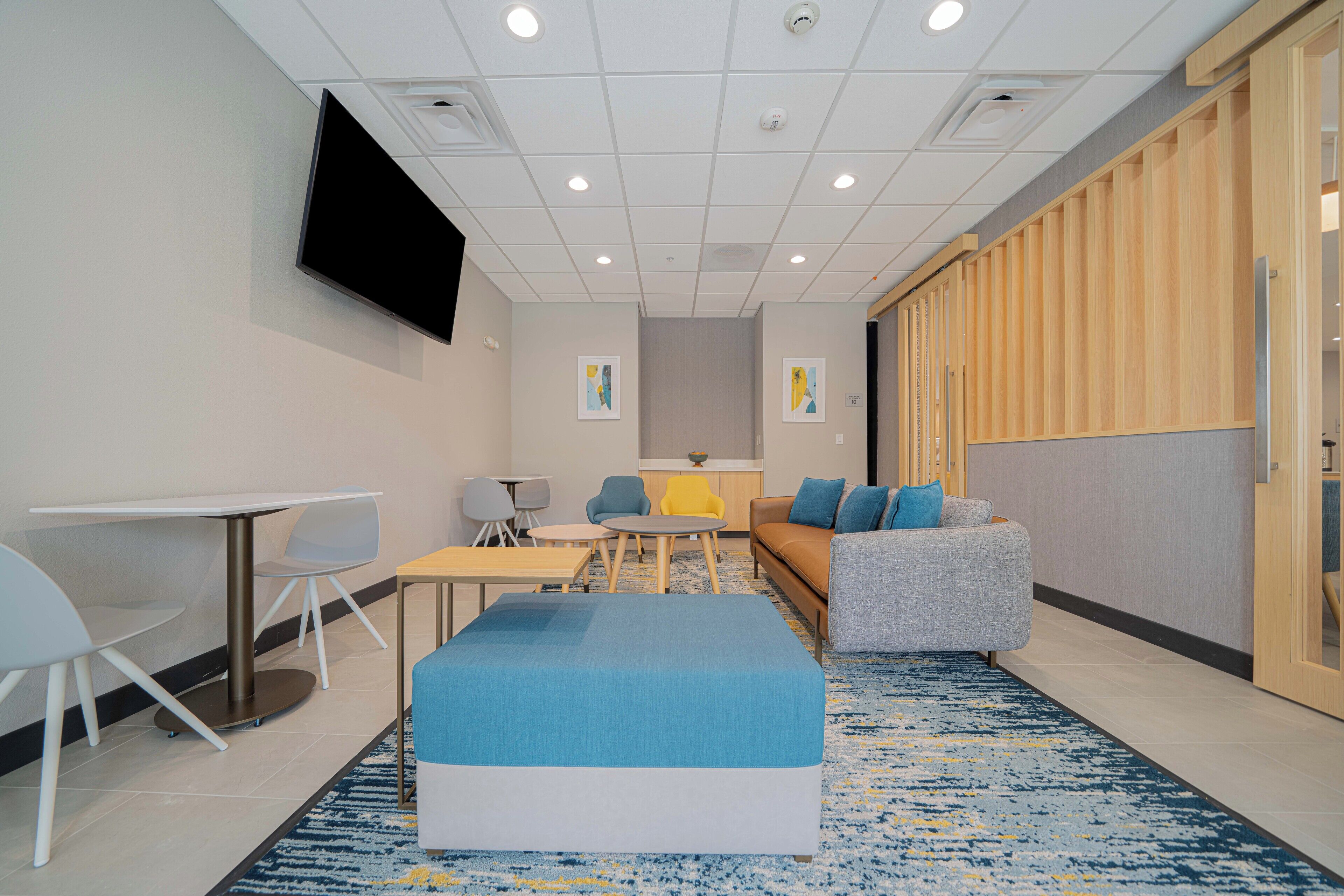 A modern hotel room with a flat-screen TV, a sofa, a small table, and a blue ottoman.