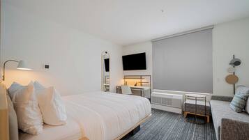 Quarto de hotel com uma cama grande, uma escrivaninha, uma cadeira, uma mesinha, um sofá, uma televisão e uma janela com cortina.
